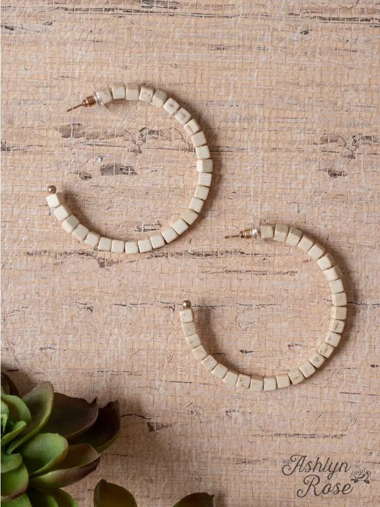 Cream-colored square beaded hoop earrings displayed on a textured neutral fabric background with green succulent accents.