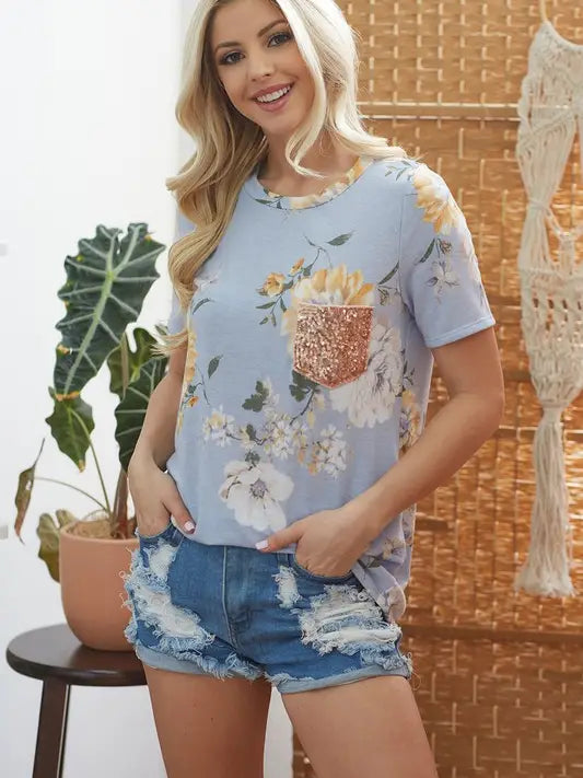 Woman modeling a light blue floral print t-shirt with rose gold sequin pocket and distressed denim shorts in a boho home setting with plants and woven decor.