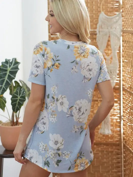 Back view of woman wearing light blue floral t-shirt with yellow and white flowers, in a boho-style room with macrame wall hanging and indoor plants.