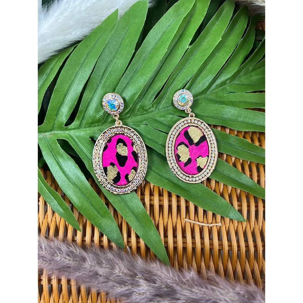 Boots & Bling Earrings featuring oval-shaped fuchsia leopard print centers framed in gold with rhinestone detailing, topped with iridescent stud posts, displayed on green palm leaves over a wicker surface.
