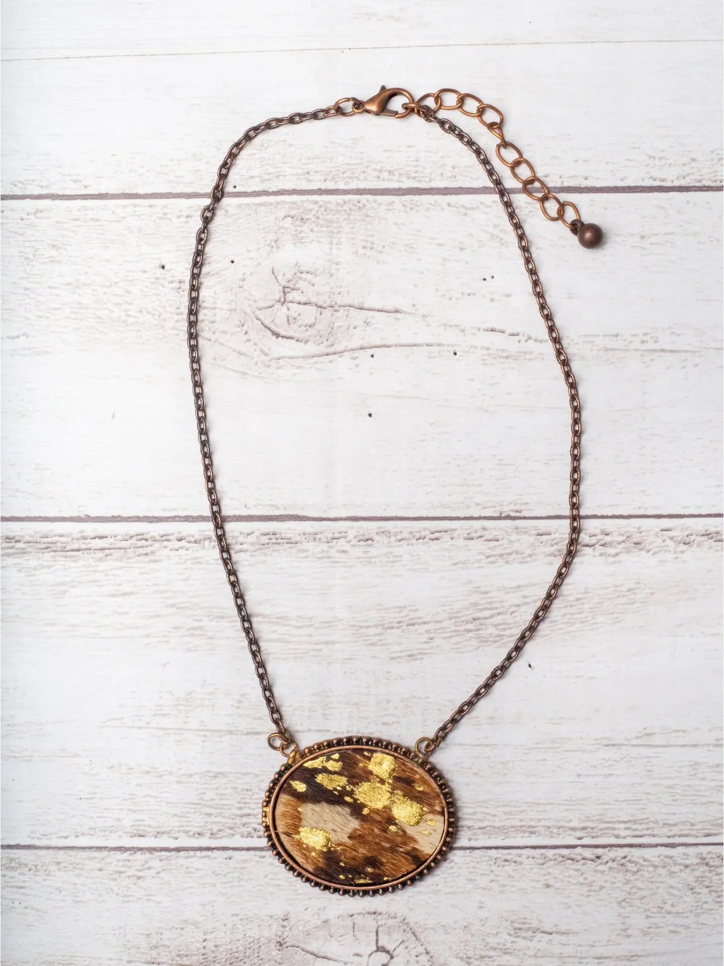 Oval cowhide pendant necklace with gold foil accents and a bronze chain, displayed on a light wood background.