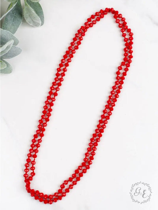 Long double-strand necklace made of bright red faceted beads, styled in an oval shape on a white background with green leafy accents in the corner.