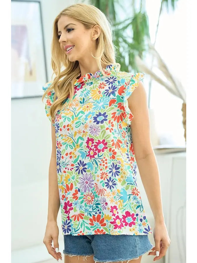 Woman in a sleeveless floral print ruffle top with mock neck, featuring colorful flowers in pink, orange, and blue, paired with denim cutoff shorts, posing indoors with modern decor.