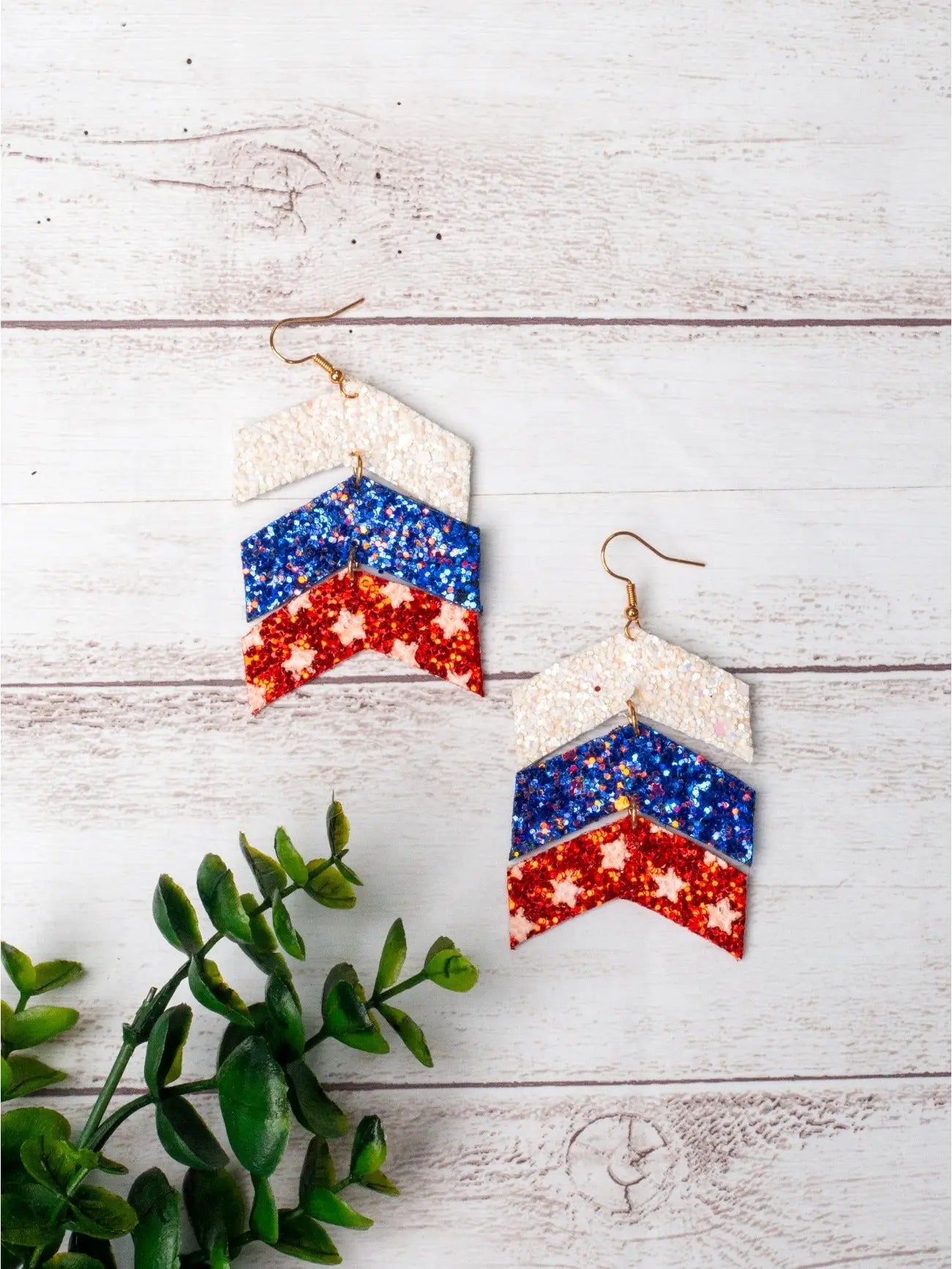 Firecracker Chevron Drops — a pair of red, white, and blue glitter tiered chevron earrings with gold hooks — displayed on a light wood background with green foliage accent.