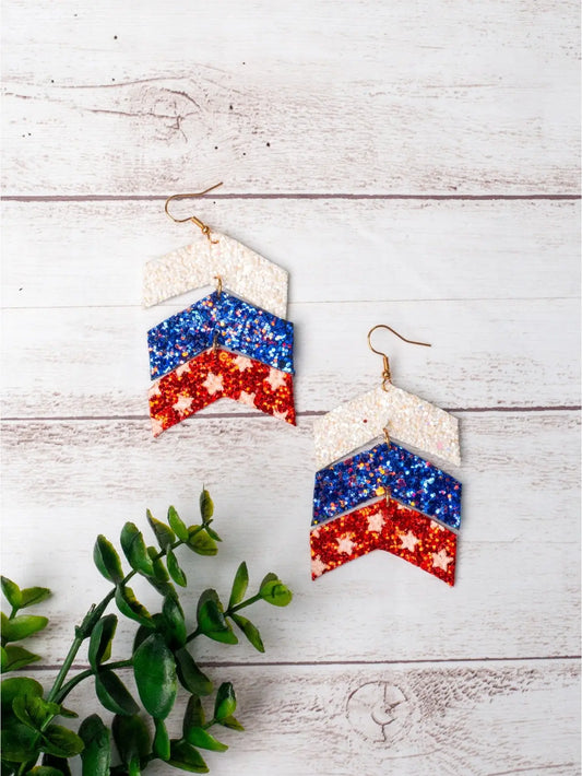 Firecracker Chevron Drops — a pair of red, white, and blue glitter tiered chevron earrings with gold hooks — displayed on a light wood background with green foliage accent.