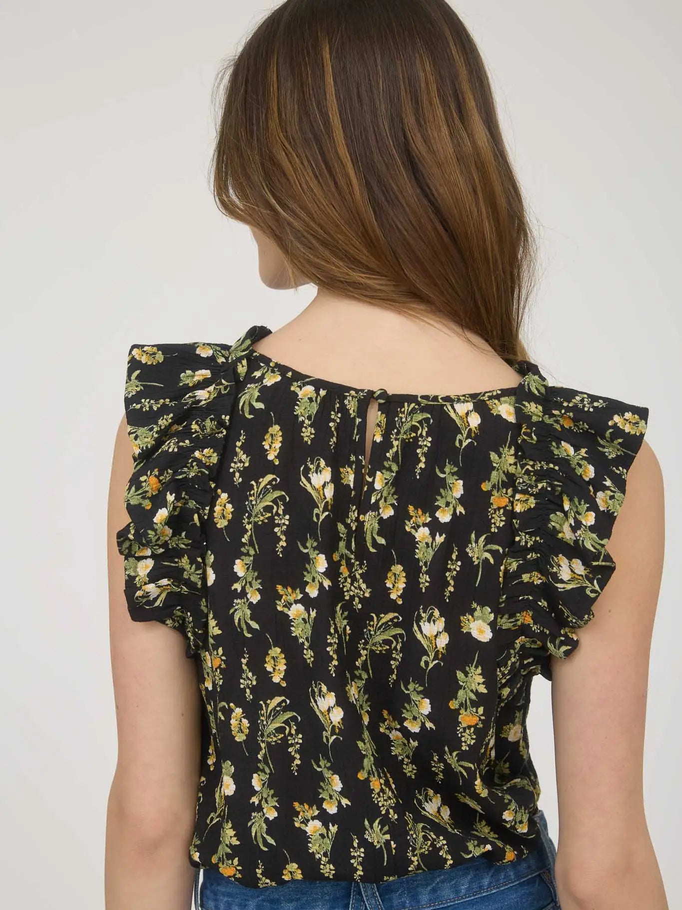 Back view of women's sleeveless black woven blouse with yellow and white floral print, round neckline, ruffle sleeve detail, and back keyhole eye closure made from rayon-blend fabric