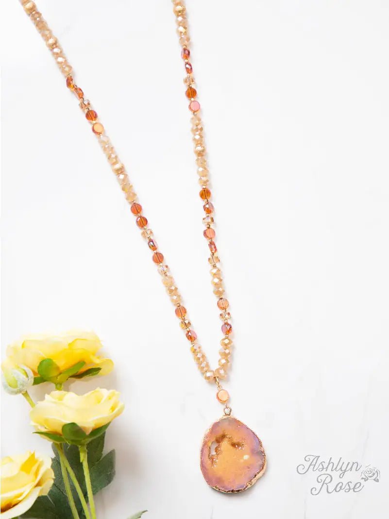 Beaded necklace in warm amber and peach tones with a shimmering druzy geode pendant, styled on a white background with yellow roses.