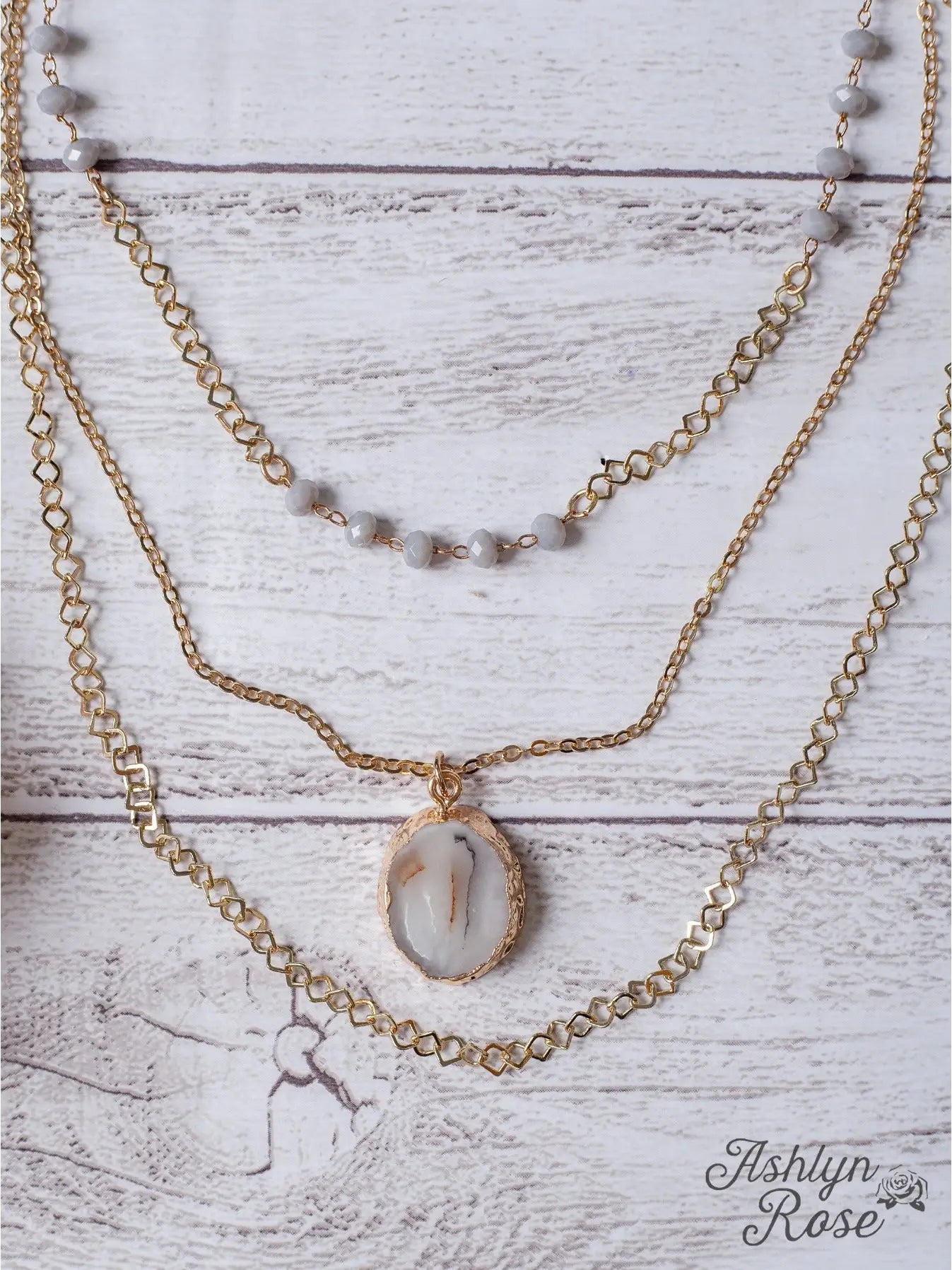 Triple-layered gold necklace with gray beaded detailing and a natural stone pendant centerpiece, displayed on a whitewashed wood background with "Ashlyn Rose" branding in the corner.
