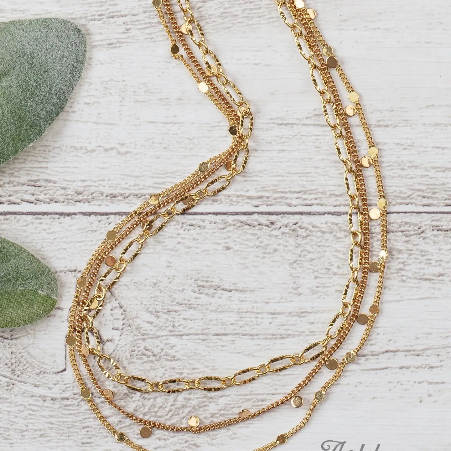 Triple-layered gold necklace with mixed chain styles and small shiny disc accents, displayed on a white wooden background next to green leaves.