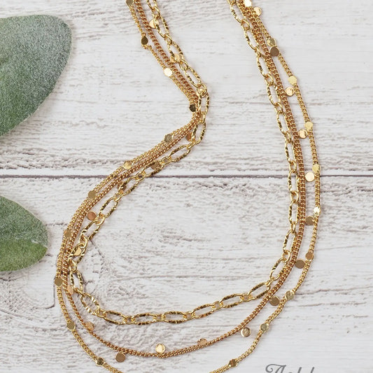 Triple-layered gold necklace with mixed chain styles and small shiny disc accents, displayed on a white wooden background next to green leaves.