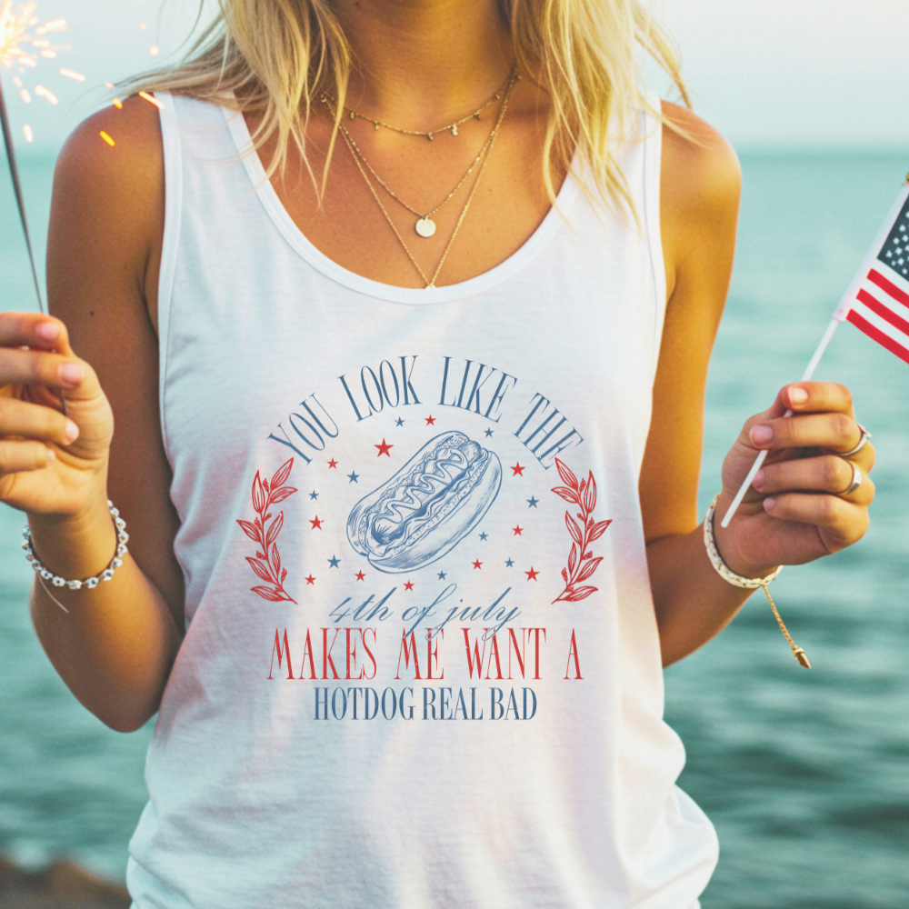 Woman wearing a white racerback tank top with a patriotic graphic that reads, “You look like the 4th of July, makes me want a hotdog real bad,” accompanied by red and blue stars, laurel graphics, and a sketch of a hot dog. She is holding a lit sparkler in one hand and a small American flag in the other, standing by the water during sunset.