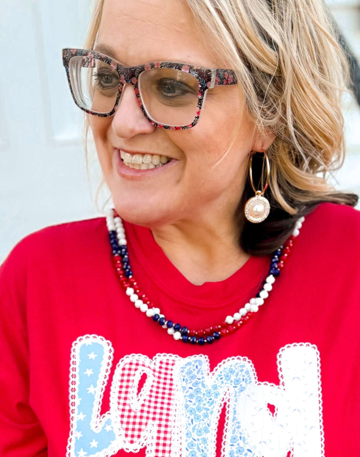Close-up of a woman wearing a red shirt with patriotic lettering and a layered beaded necklace in red, white, and blue. The necklace features alternating sections of small, round beads, capturing a festive Americana theme perfect for patriotic holidays.