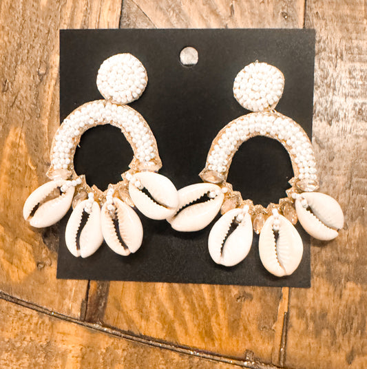 Statement earrings with beaded circular tops and semi-hoop shapes adorned with white cowrie shells, displayed on a black card atop a rustic wooden surface. Coastal and boho-inspired design.