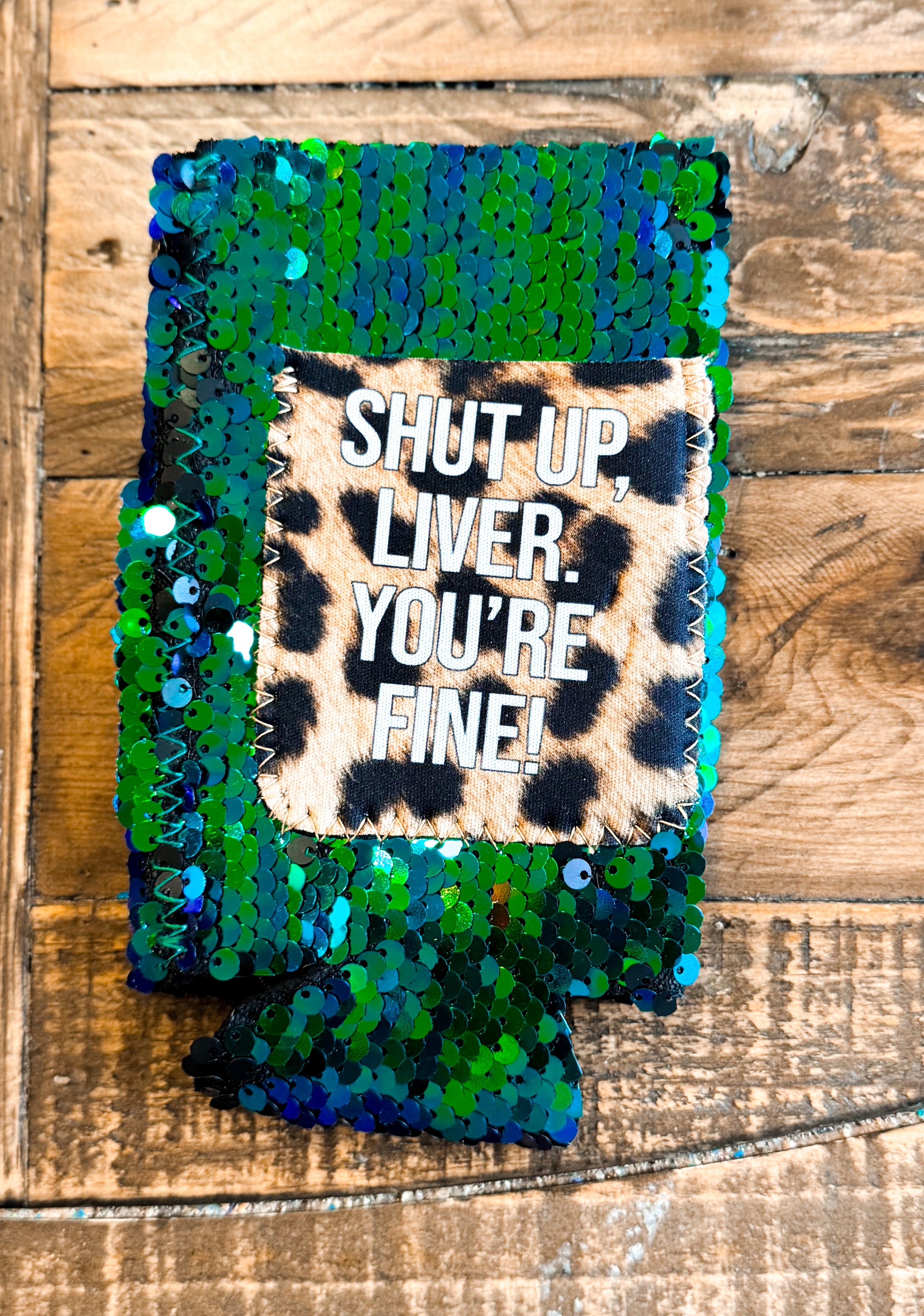 Green and blue sequin slim can cooler with a leopard print patch that reads “SHUT UP, LIVER. YOU’RE FINE!” in white block letters.