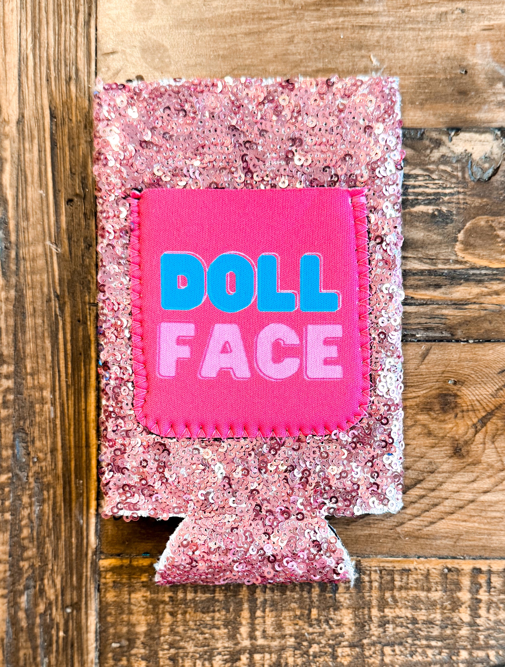 Light pink sequin slim can cooler with a hot pink patch that says “DOLL FACE” in bold blue and pastel pink block letters.