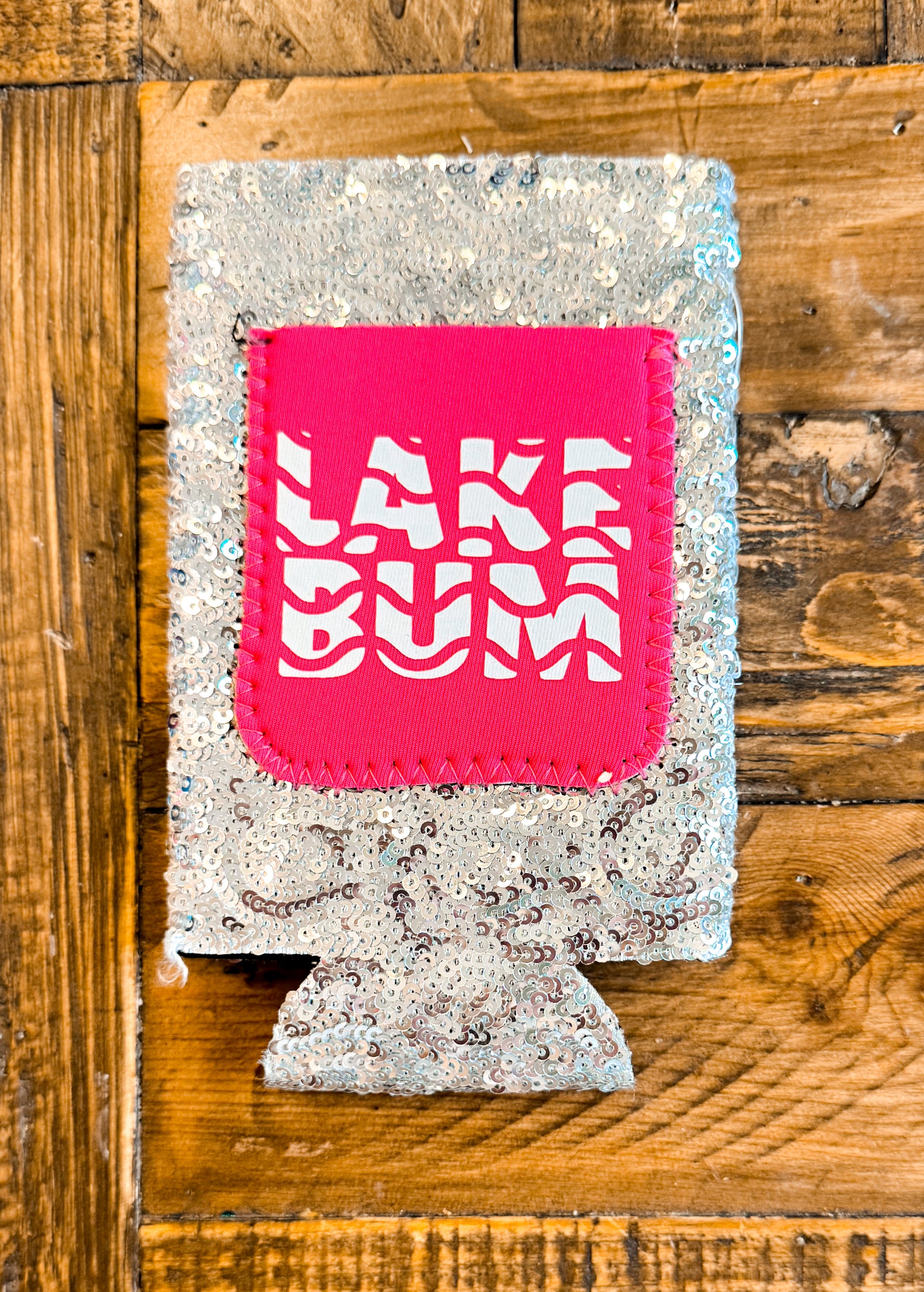 Silver sequin slim can cooler with a bright pink patch that says “LAKE BUM” in white wavy block lettering.