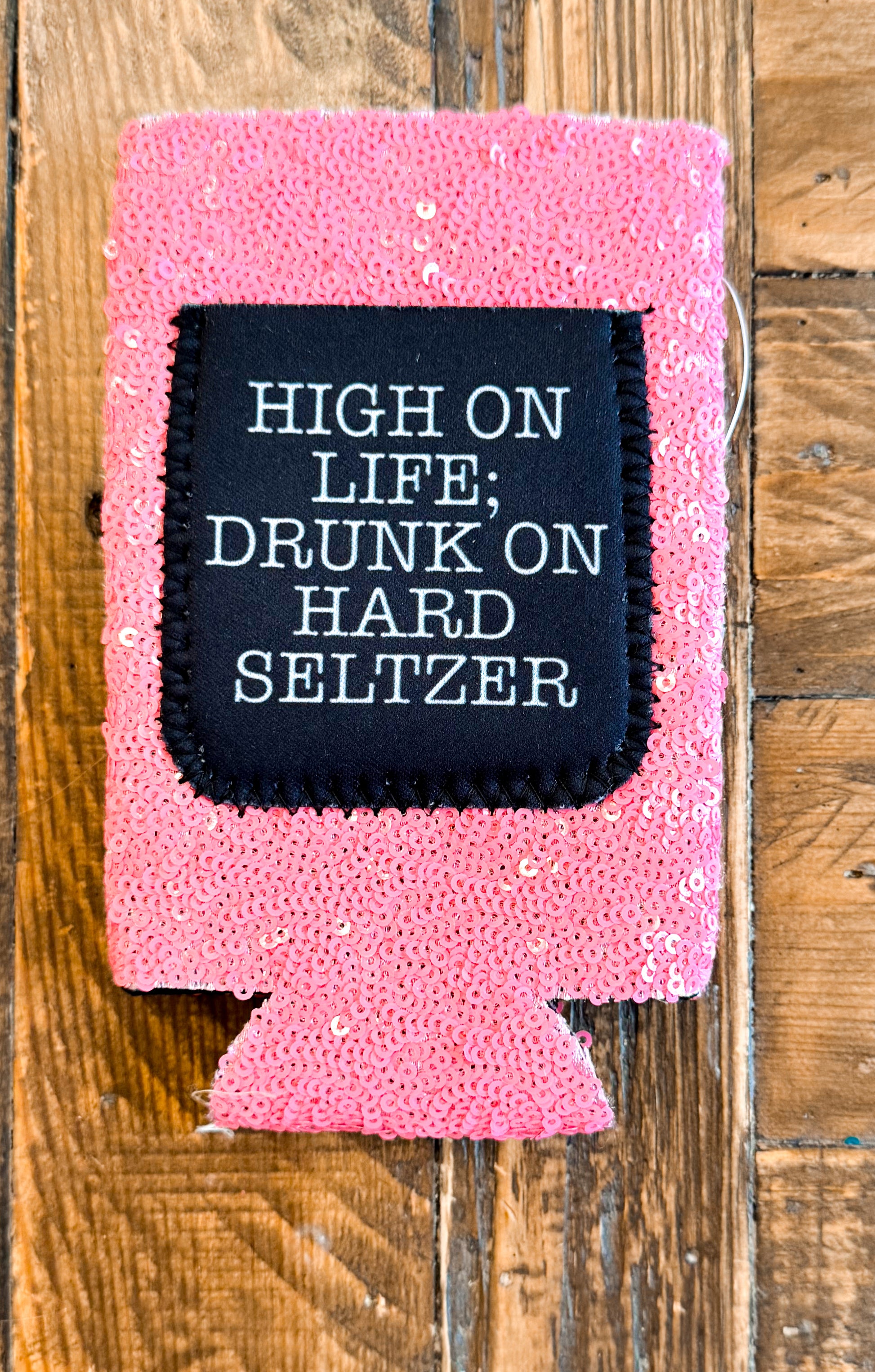 Pink sequin slim can cooler with a black patch that says “HIGH ON LIFE; DRUNK ON HARD SELTZER” in white typewriter-style font.