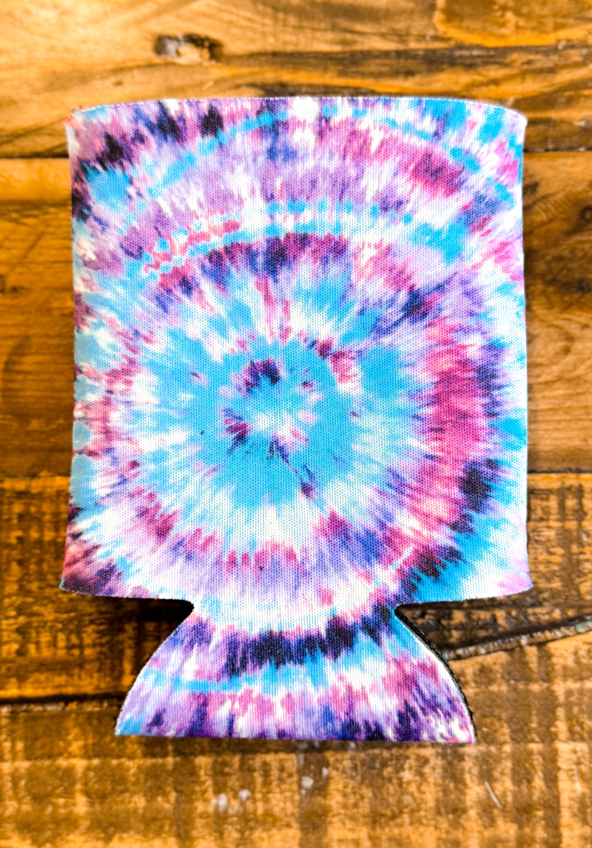 Tie-dye can cooler featuring a vibrant spiral design in shades of turquoise, lavender, magenta, and white, displayed on a rustic wooden background.