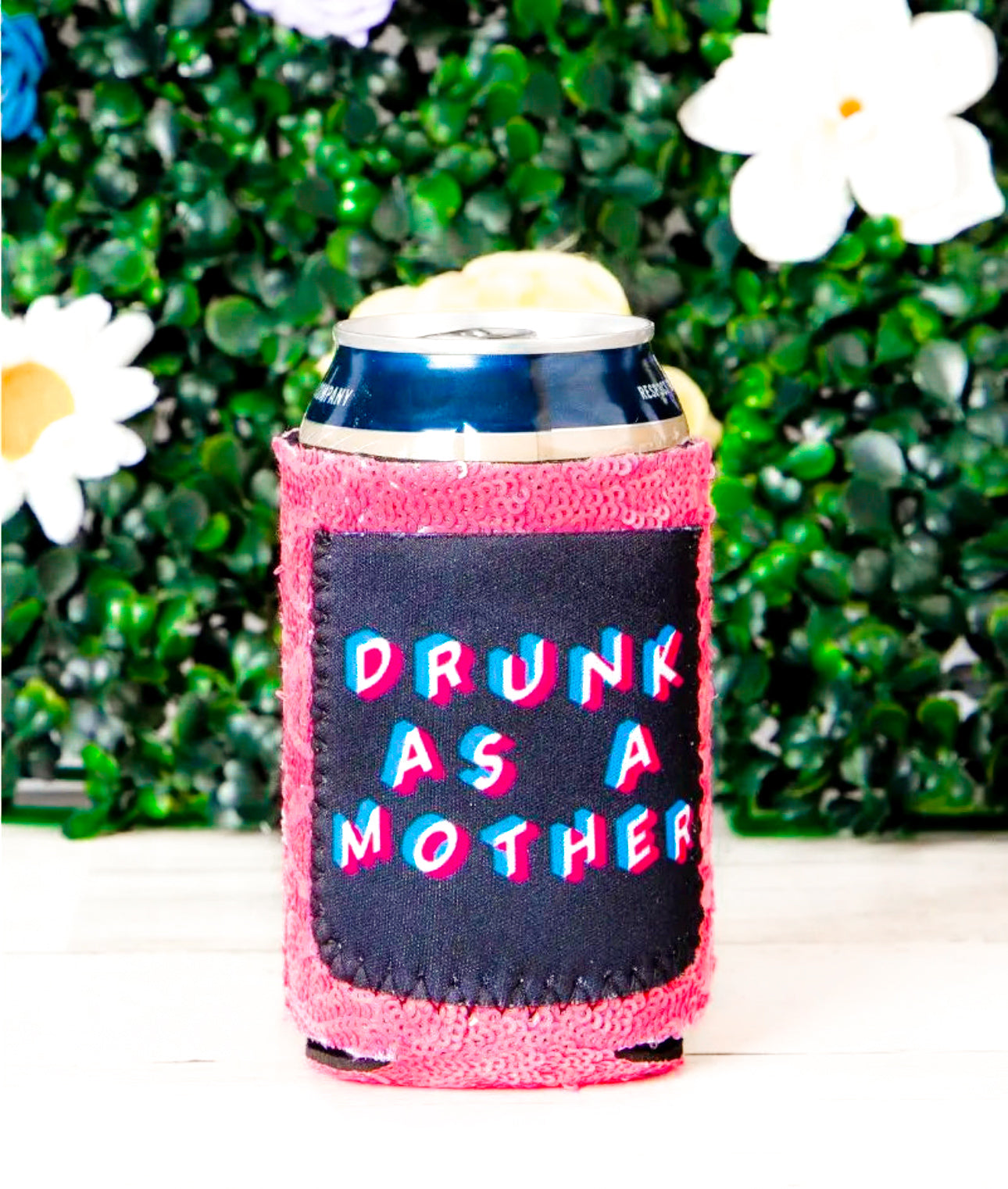 Pink sequin can cooler with a black patch that reads âDRUNK AS A MOTHERâ in bold, 3D-style red, white, and blue text. A can is placed inside, and the background features green artificial foliage with white and purple flowers.