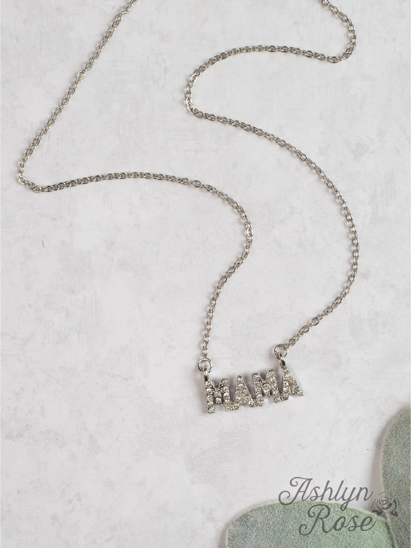 Silver chain necklace with a rhinestone-encrusted "MAMA" pendant displayed on a light textured surface, with "Ashlyn Rose" branding in the corner.