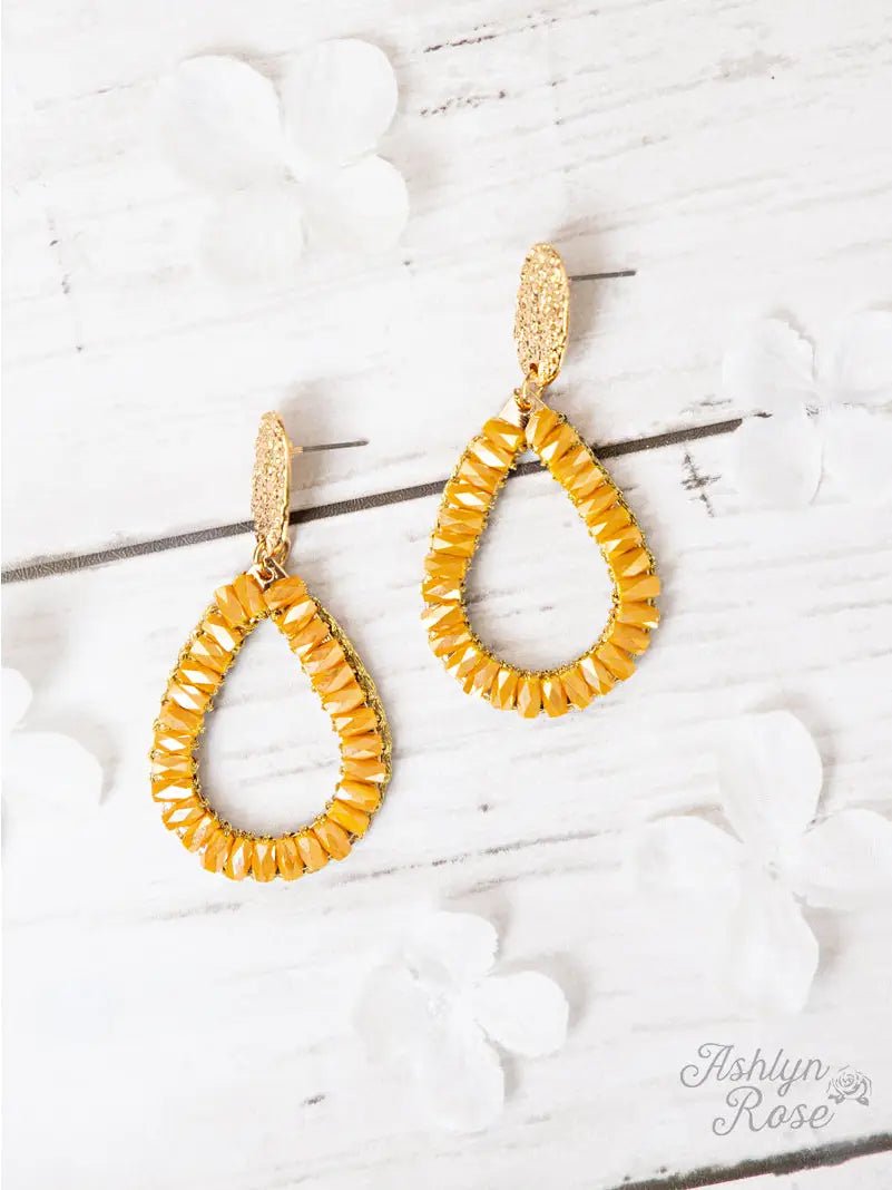 Teardrop-shaped earrings wrapped in mustard yellow beads with textured gold posts, displayed on a white wooden background with white flower accents.