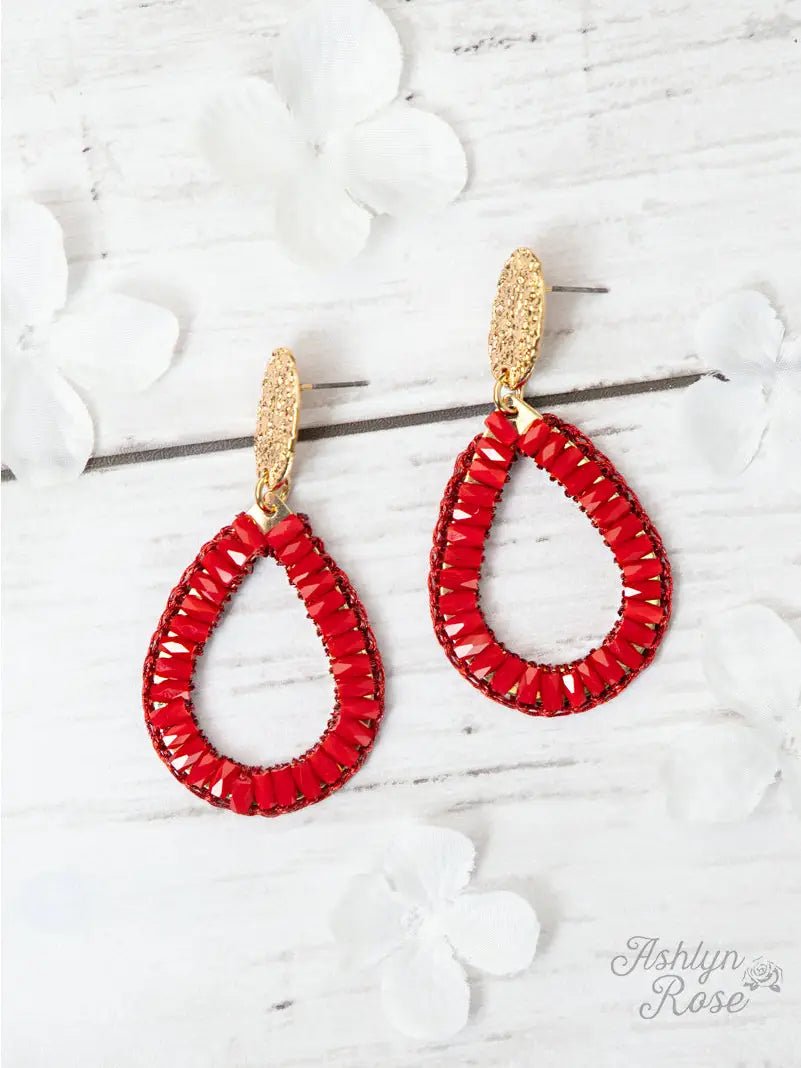 Teardrop-shaped earrings wrapped in red beads with textured gold posts, displayed on a white wooden background with white flower accents.