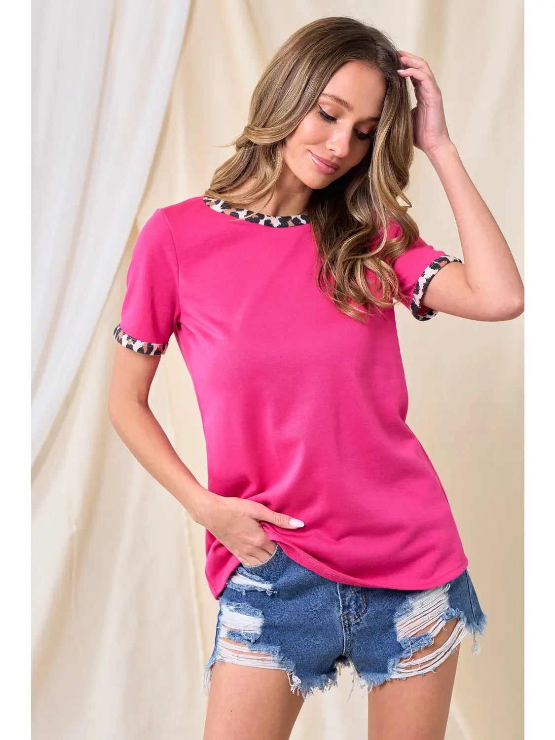 Woman modeling a bright pink short sleeve t-shirt with leopard print trim on collar and cuffs, paired with high-rise distressed denim shorts, standing against a neutral backdrop.