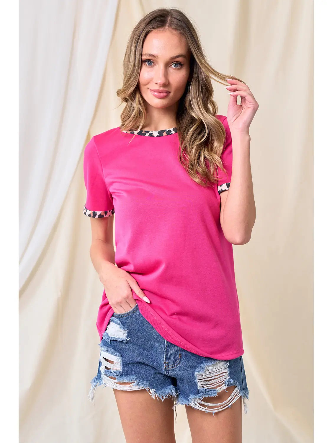 Woman wearing a hot pink t-shirt with leopard print neckline and sleeve cuffs, styled with distressed high-rise denim shorts, standing against a light fabric backdrop.