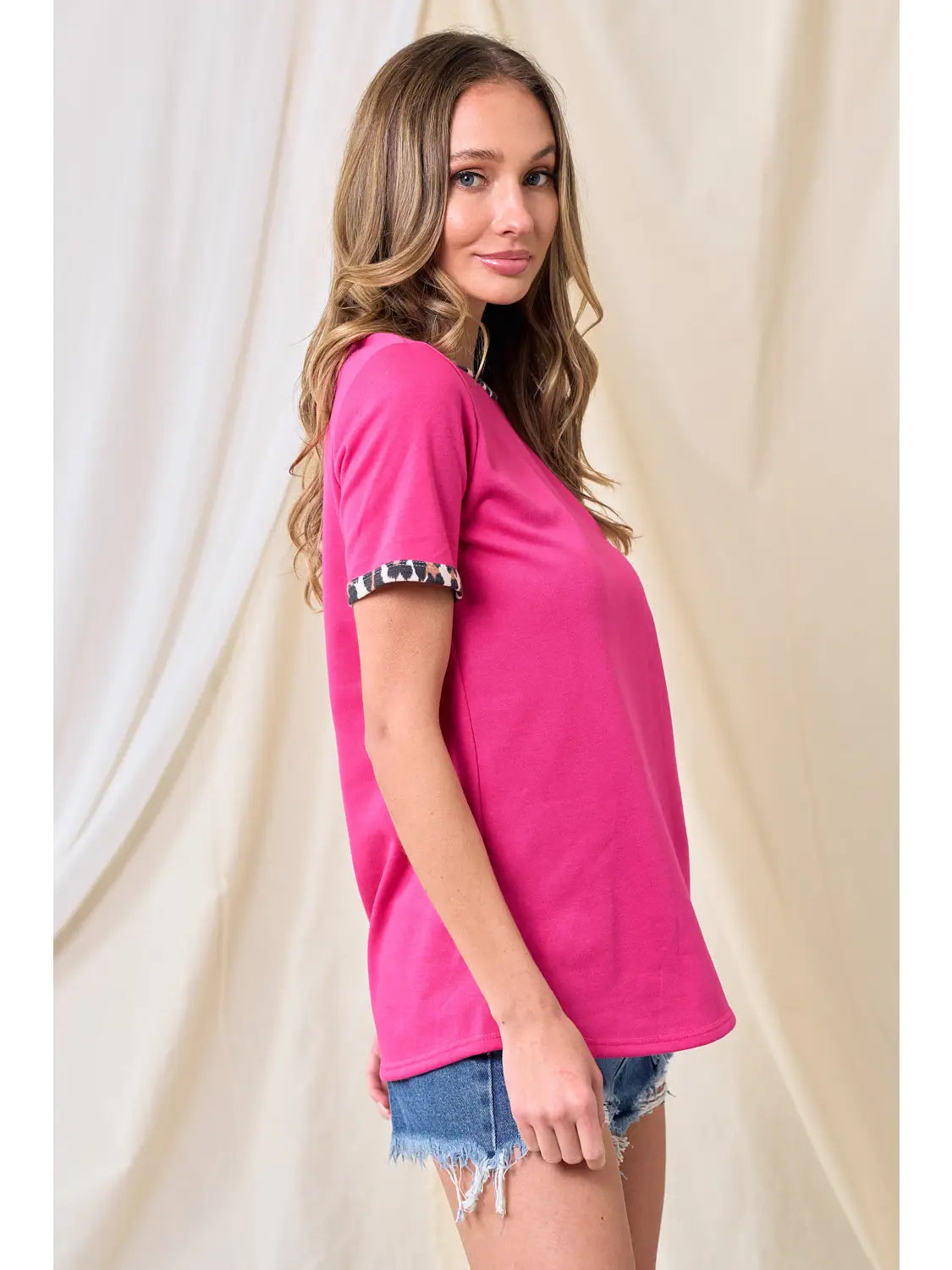 Side view of woman wearing a hot pink t-shirt with leopard print sleeve cuffs and distressed denim shorts, standing in front of a neutral fabric backdrop.
