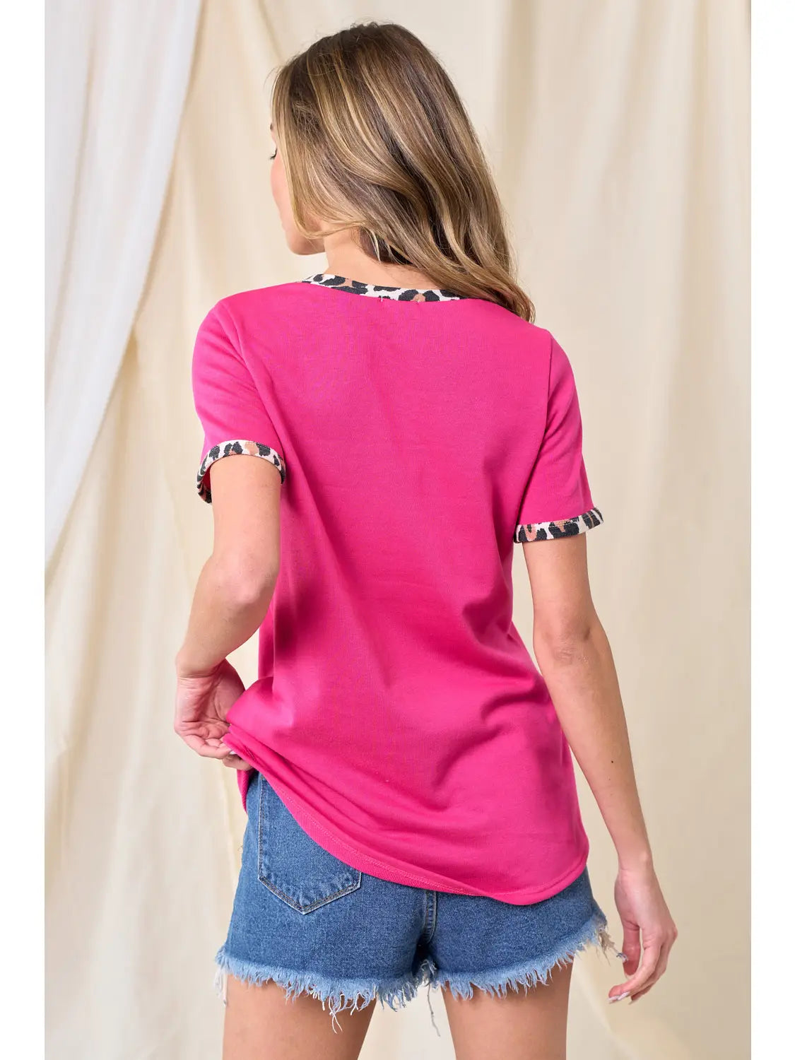 Back view of woman wearing a hot pink t-shirt with leopard print trim on neckline and sleeves, paired with frayed hem denim shorts, standing in front of a neutral backdrop.