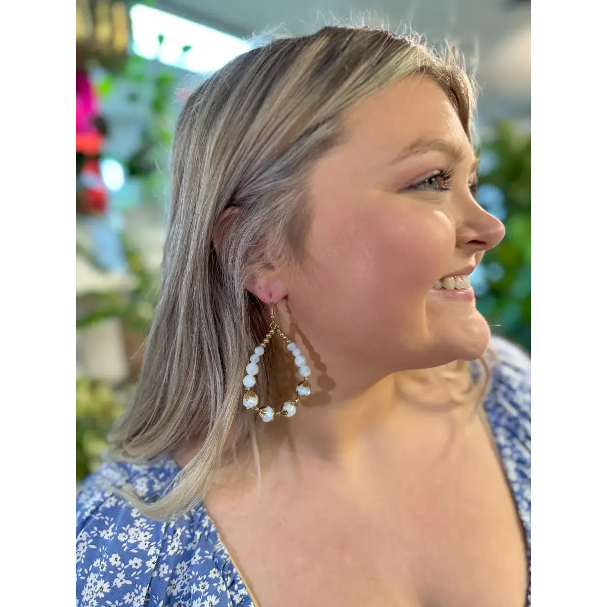 Woman smiling and wearing large teardrop-shaped earrings with white beads and gold wire, styled with a light blue floral dress.