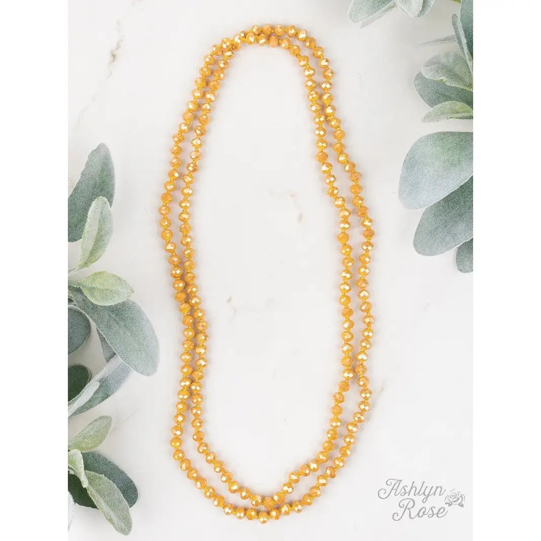 Desert Marigold Necklace featuring mustard yellow faceted beads, styled as a long double-wrap strand, displayed on a white marble surface with soft green foliage accents.