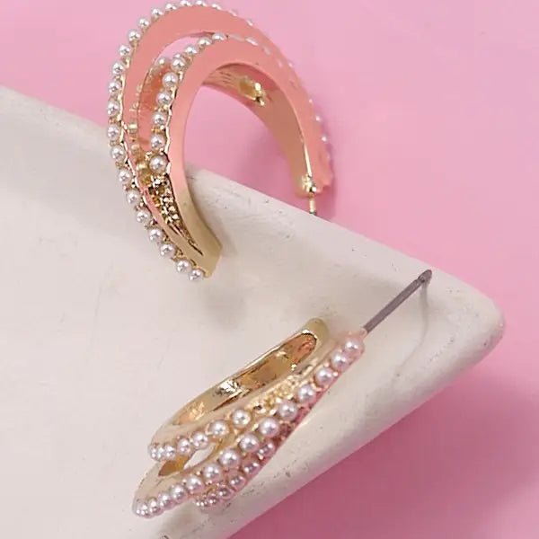 Gold triple-layered hoop earrings embellished with small faux pearls, displayed on a white dish with a pink background.