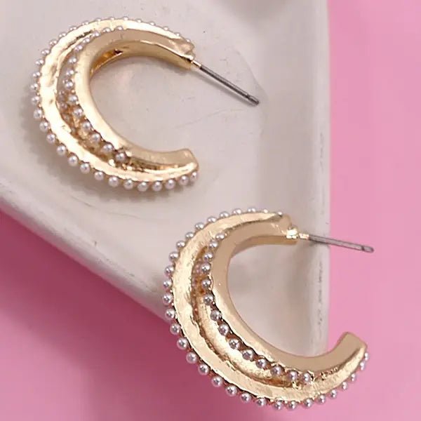 Gold triple-layered hoop earrings embellished with small faux pearls, displayed on a white dish with a pink background.