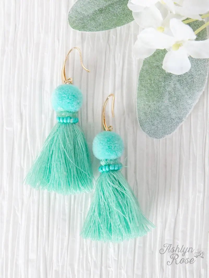 A pair of mint green tassel earrings with pom-pom tops, delicate bead detailing, and gold-tone hooks, displayed on a textured white background with greenery and white flowers nearby.