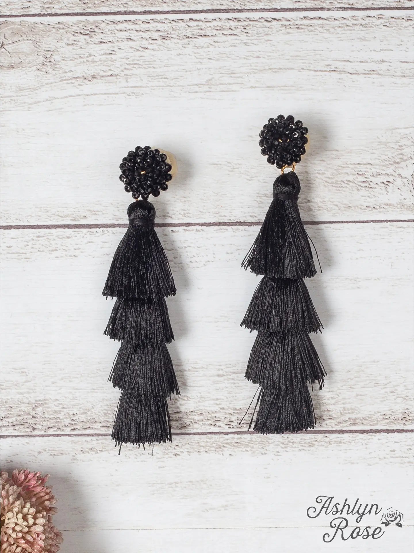 Pair of black tiered tassel earrings with beaded stud tops, displayed on a light wood background with a small pink flower in the corner.