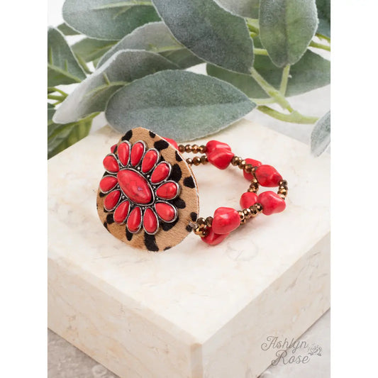 Run Wild Bracelet featuring a leopard-print focal with red floral stone design, bronze faceted beads, and chunky red stone accents, displayed on a marble platform with soft green eucalyptus in the background.