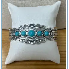 Silver cuff bracelet with intricate etching and five turquoise stones displayed on a white cushion.