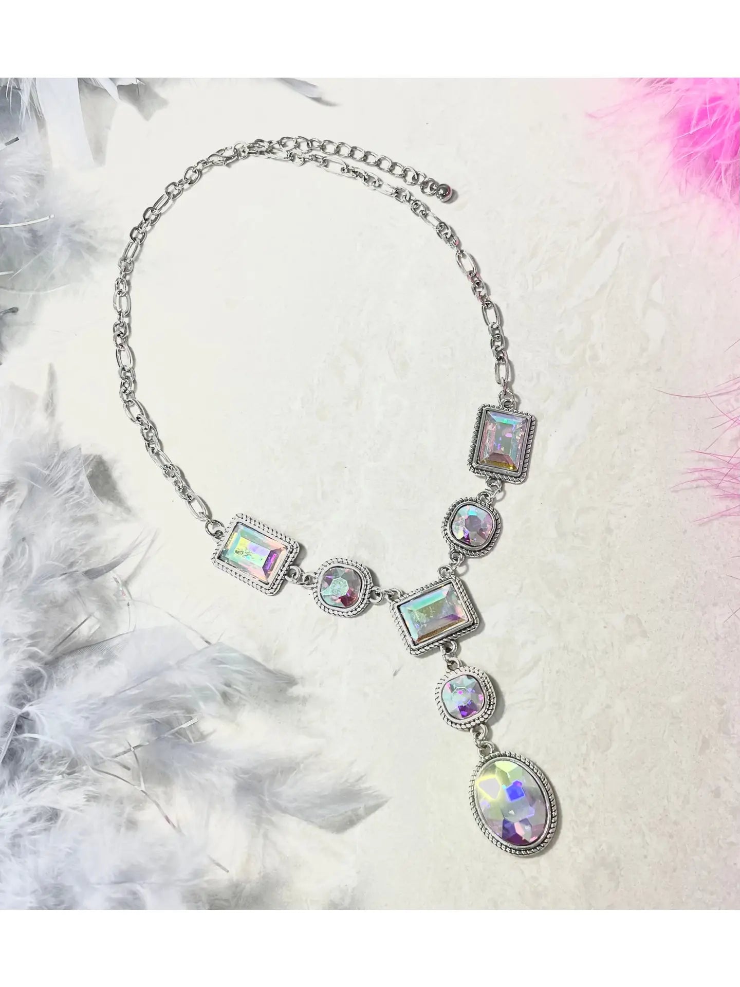 Silver statement necklace featuring geometric iridescent crystals in rectangular, round, and oval shapes, arranged in a cascading design on a chunky silver chain, displayed against a soft cream background with white and pink feathers.