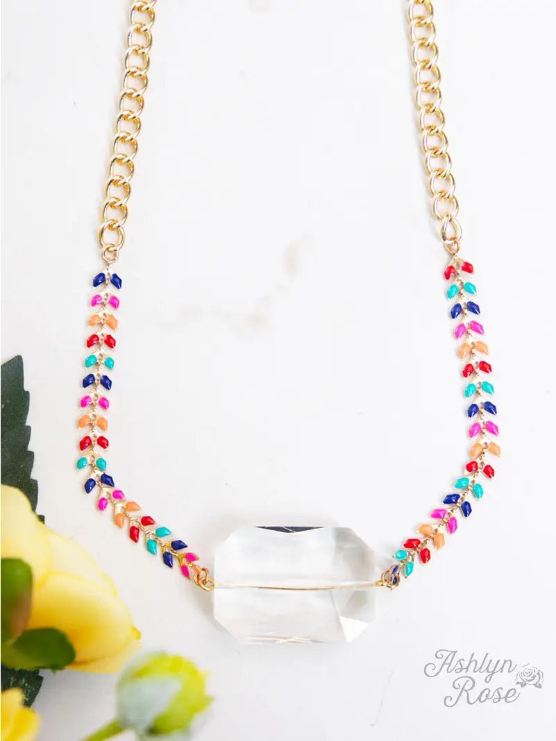 Colorful enamel chain necklace with a large clear faceted glass stone pendant, accented by gold links, displayed on a white background with yellow flowers in the corner.