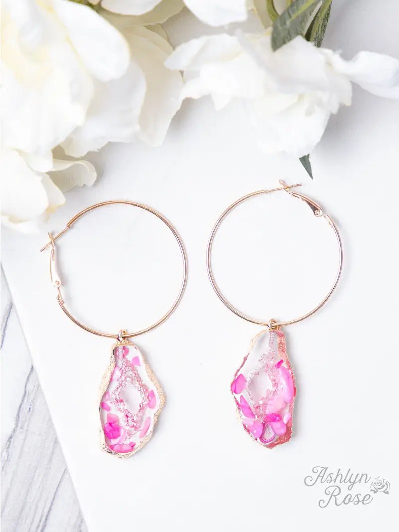 Gold hoop earrings with dangling irregular pink and clear geode-style resin pendants outlined in gold, displayed on a white surface with soft white flowers in the background.
