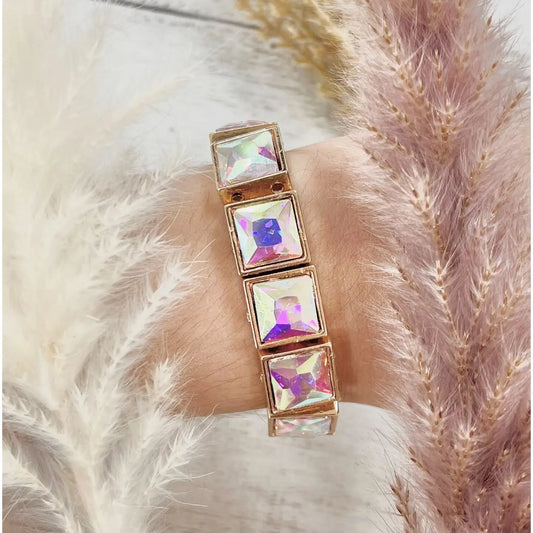 Close-up of the Prism Pop Bracelet — a gold-toned stretch bracelet with large iridescent square gemstones — worn on wrist and surrounded by soft blush and white pampas grass.