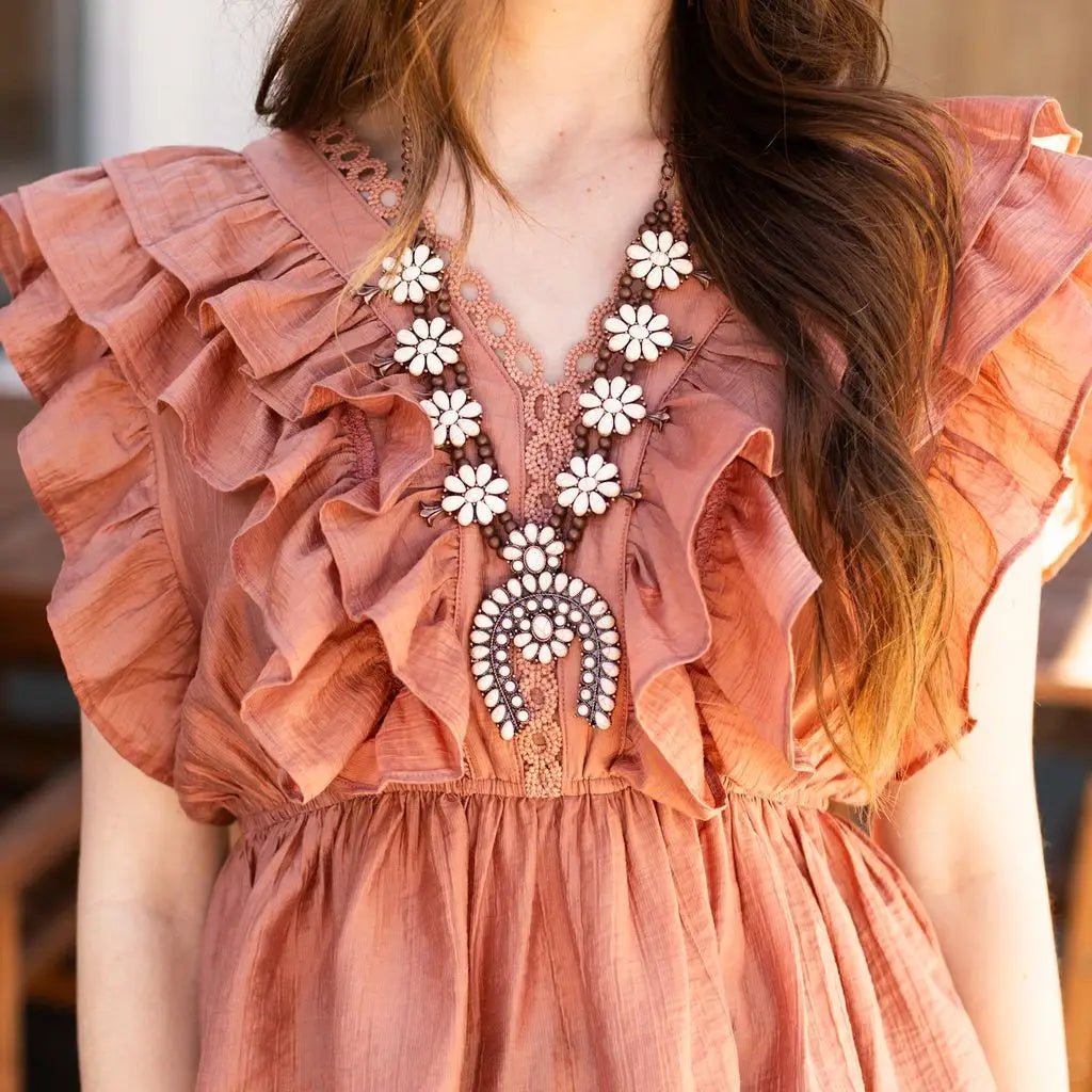 Woman wearing the Sierra Bloom Necklace — featuring cream floral clusters and a bronze squash blossom pendant — paired with matching earrings, a ruffled terracotta blouse, and denim bottoms.