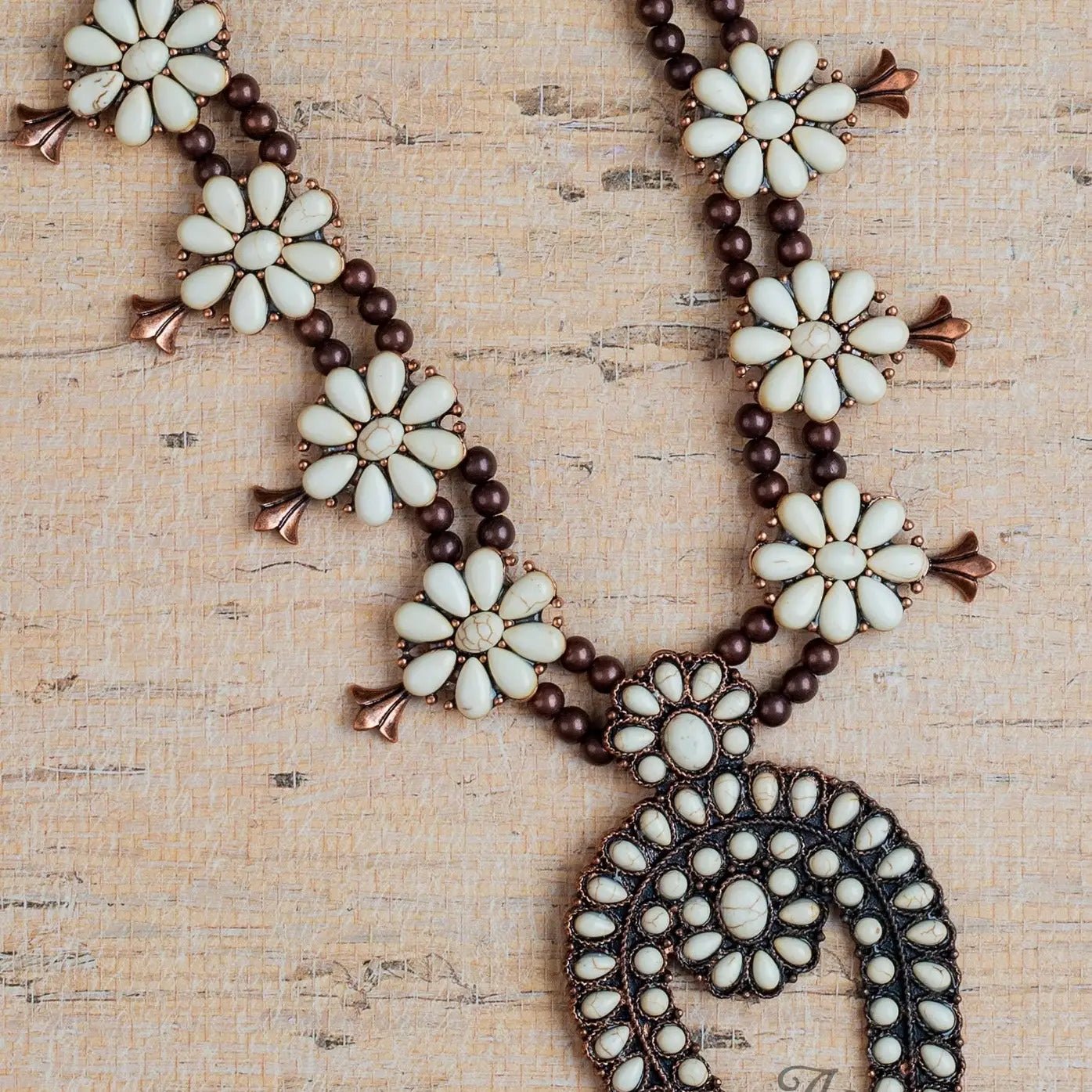 Sierra Bloom Necklace featuring cream floral bead clusters and a bold squash blossom pendant, accented with bronze beads and metallic details, displayed on a textured beige background with "Ashlyn Rose" watermark.