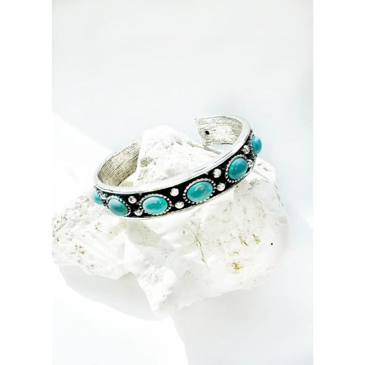 A silver open cuff bracelet with oval turquoise stones and vintage-inspired detailing, styled on a white stone background with bright lighting.