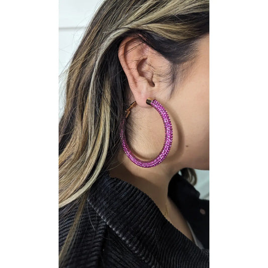 Close-up of a woman wearing the Pink Power Hoops — large hoop earrings covered in sparkling hot pink crystals — styled with straight, highlighted hair and a textured black top.