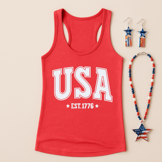 Flat lay image of a red heathered racerback tank top with bold white text reading “USA EST. 1776” printed on the front. The tank is paired with patriotic accessories including star-shaped red, white, and blue earrings and a beaded necklace featuring a star pendant with the American flag design. The background is a soft neutral beige.