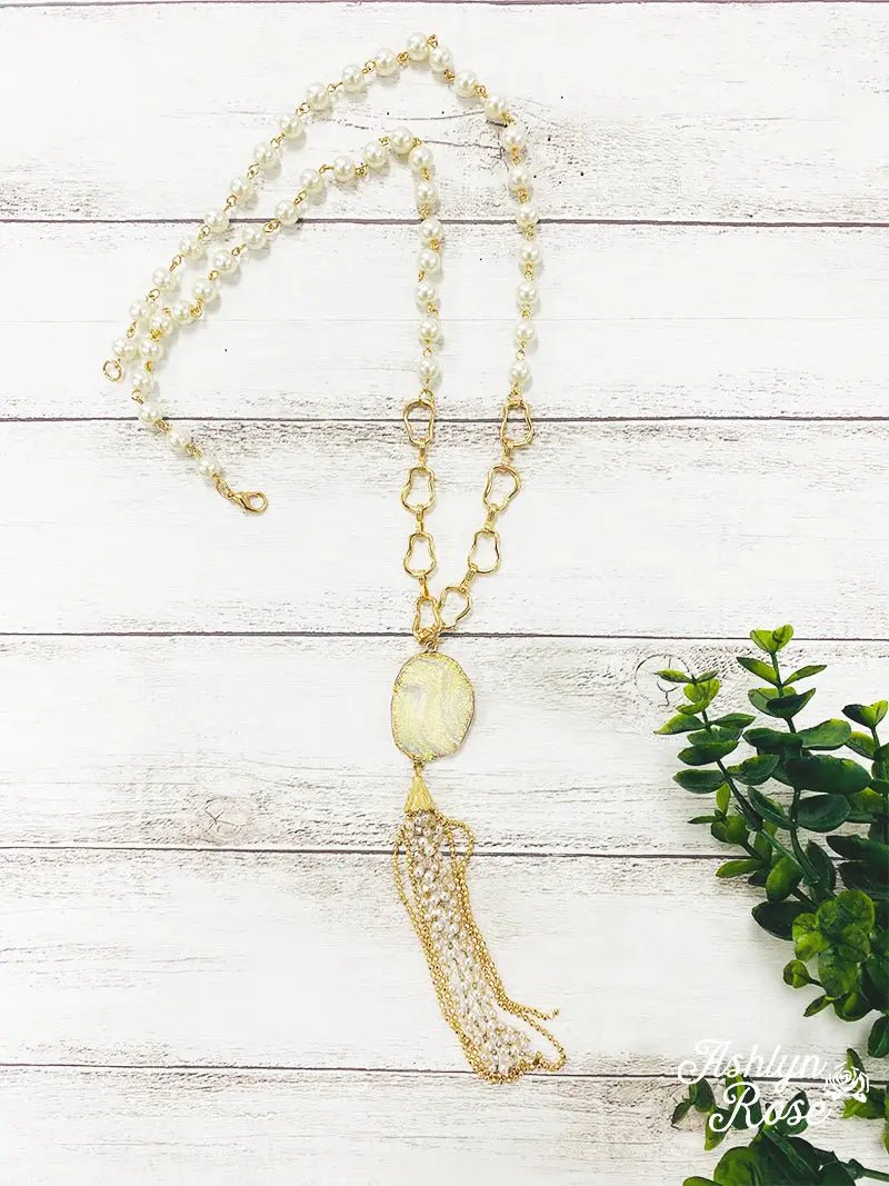 Long beaded necklace featuring pearl-like beads, hammered gold links, a large iridescent stone pendant, and cascading tassels made of gold chains and tiny pearls, styled on a white wood background with greenery.