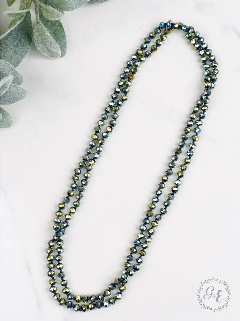 Double-strand beaded necklace with metallic teal, gold, and green faceted beads arranged in a loop on a white surface with soft greenery in the corner.