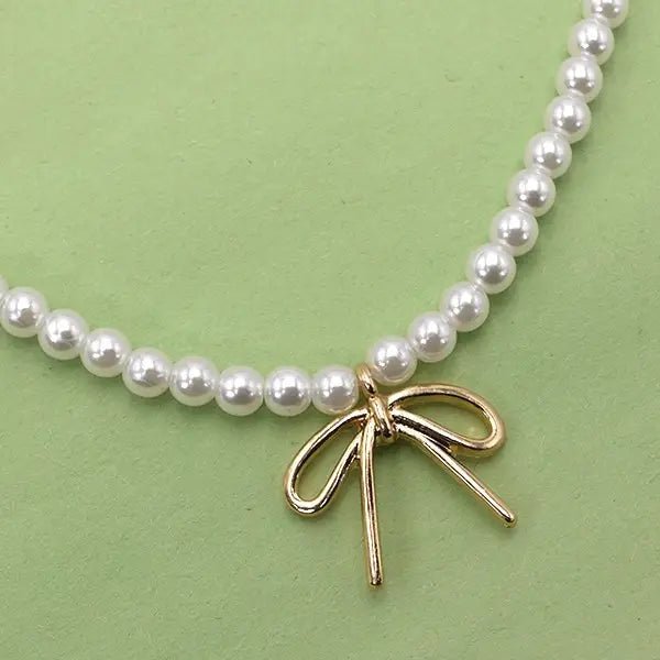 Close-up of a pearl beaded necklace with a small gold-tone bow pendant, displayed on a light green background.